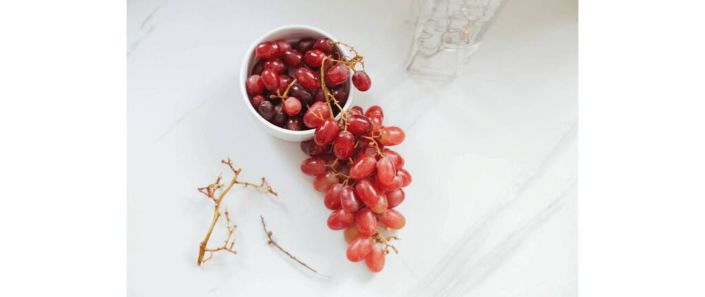 sobre uma mesa branca 1 pote cheio de uva vermelha, 1 cacho de uva vermelha e 1 galho sem uvas