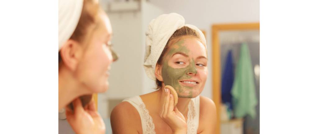 Mulher loira de toalha na cabeça se olhando no espelho para tirar a máscara facial de argila verde