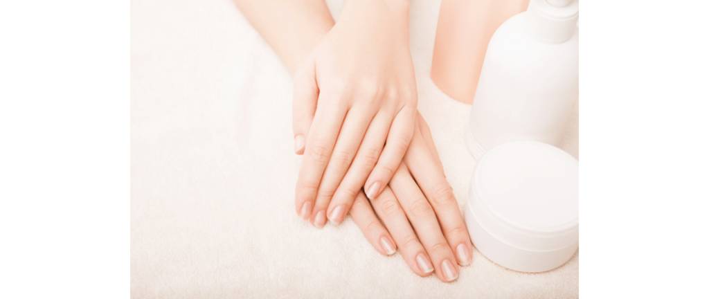 mão feminina direita sobre a mão esquerda, mostrando bem as unhas saudáveis, ao lado potes de produtos para unhas e mãos